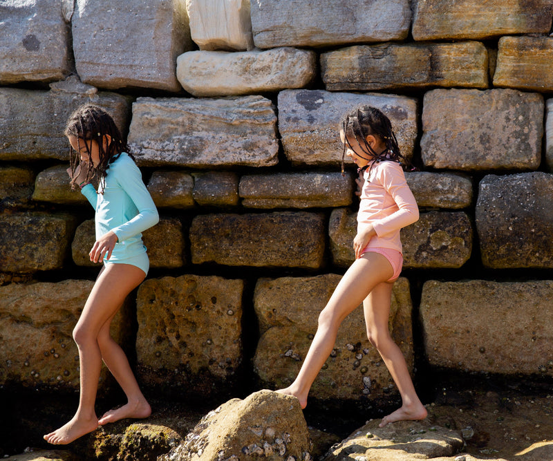 Two Girls Walking On Rocks Wearing Green And Peach Rash Guard And SwimBrief 