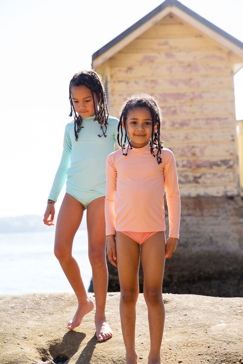Two Girls Standing On Rock Wearing Rose Peachie And Green Rash Guard And Swim Brief 