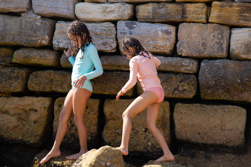 Two Girls Climbing Over Rocks Wearing Rose Peachie And Green Rash Guard And Swim Brief 